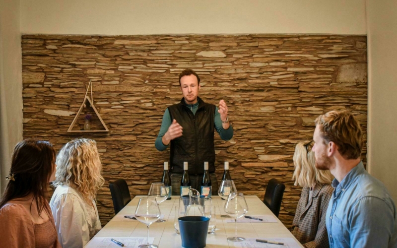 Photo of a man in front of wine bottles talking to a group of people sitting at a white table