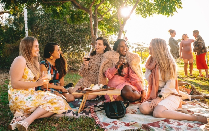 girls sitting under a tree on picnic blankets. Drinking wine and laughing.