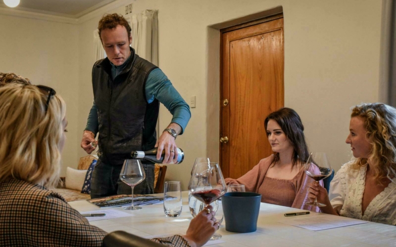 A man pouring wine into a glass. Three women are sitting at a white wine tasting table.