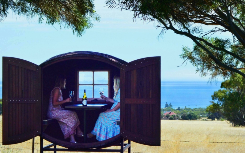 Two ladies drinking wine inside the wine barrel tasting pod over looking sea views
