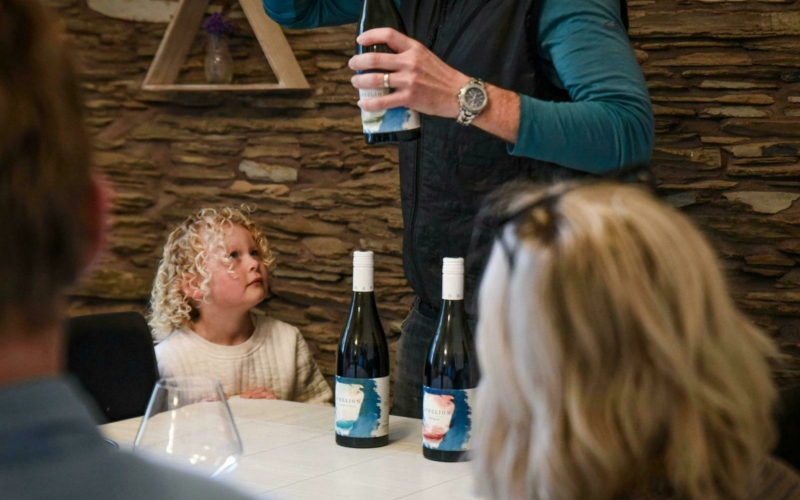 A curly haired girl looking at her dad who is pouring wine for some women sitting at a white table